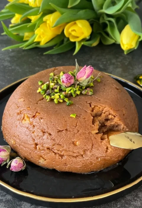 Semolina halva moulded into a dome shape on a black plate topped with pistachios and dried rose