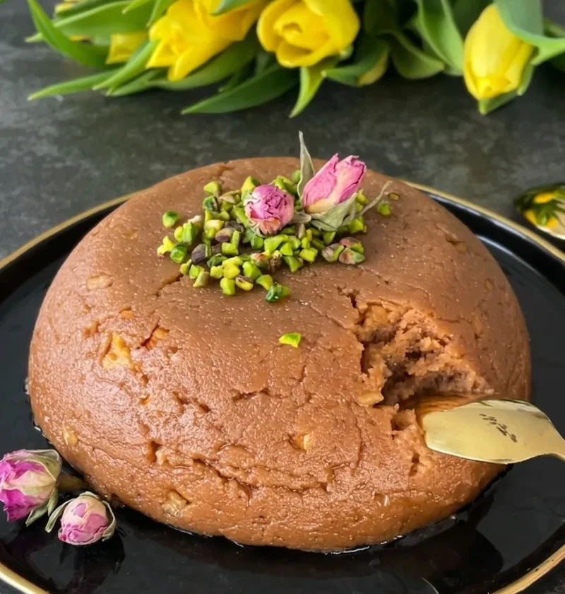 Semolina halva moulded into a dome shape on a black plate topped with pistachios and dried rose